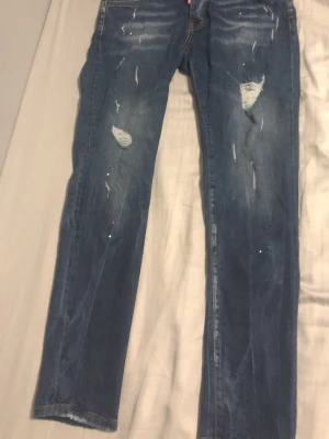 Blå slitna jeans från Dsquared2 - Säljer ett par blå jeans från Dsquared2 med distressed detaljer, slitningar och hål på benen. Jeansen har klassisk femficksmodell, raka ben och en röd logotagg vid gylfen. Perfekta för en avslappnad och trendig look.