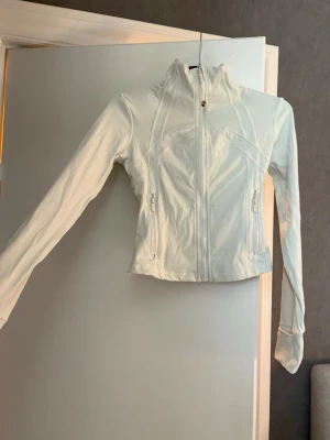 Vit träningsjacka från Lululemon - Snygg vit träningsjacka från Lululemon med figurnära passform och hög krage. Jackan har dragkedja framtill, två fickor med dragkedja och långa ärmar med tumhål. Perfekt för aktiva dagar och sportig stil. Det är den kortare modellen. Den är i storlek 2 vilket motsvarar Xs. Har bara använt 1 gång.