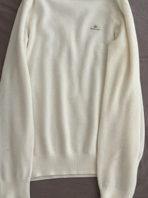 Vit stickad tröja från Gant - Klassisk vit stickad tröja från Gant med diskret logga på bröstet. Tröjan har rund halsringning, långa ärmar och ribbade muddar vid ärmslut och nederkant. Perfekt för en stilren och enkel look.