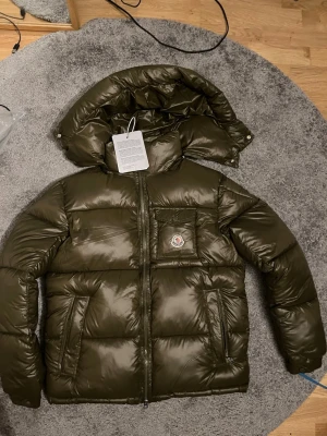 Mörkgrön dunjacka från Moncler - Snygg mörkgrön pufferjacka från Moncler med glansig finish, stor huva och dragkedja framtill. Jackan har en bröstficka med Moncler-logga och två sidofickor. Perfekt för kalla dagar och riktigt trendig look.