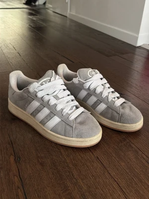 Grå Adidas Campus sneakers - Snygga grå Adidas Campus sneakers i mocka med klassiska vita ränder på sidorna och vit sula. Skorna har lågt skaft och vita skosnören. Perfekta för dig som gillar retrostil och streetwear.