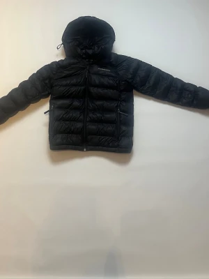 Mörkblå dunjacka från Peak Performance storlek 160 - Säljer en mörkblå dunjacka från Peak Performance med huva och dragkedja framtill. Jackan har två sidofickor med dragkedja och quiltad design som håller värmen. Perfekt för kalla vinterdagar. Jackan har storleken 160. Jackan har också en lageslapp på ena ärmen på grund av en tidigare skada på jackan, men lappe är i samma färg som jackan så det syns knappt att det är en liten del som varit sönder.