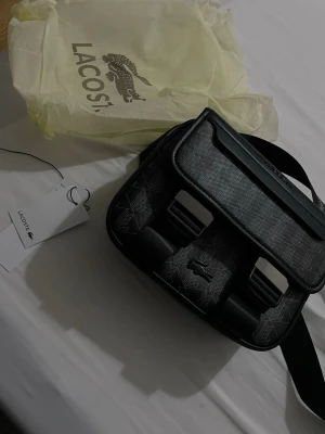  Lacoste bag - Snygg svart axelväska från Lacoste med geometriskt mönster och detaljer i konstläder. Väskan har två spännen framtill, justerbar axelrem och präglad Lacoste-logga på locket. Perfekt för dig som vill ha en stilren och modern väska.