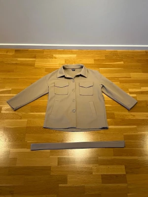 Beige overshirt från Gina Tricot - Stilren beige overshirt från Gina tricot med klassisk krage, två stora bröstfickor med lock och knäppning framtill. Tillhörande bälte för att markera midjan. Perfekt för lager-på-lager och enkel att matcha med olika outfits.