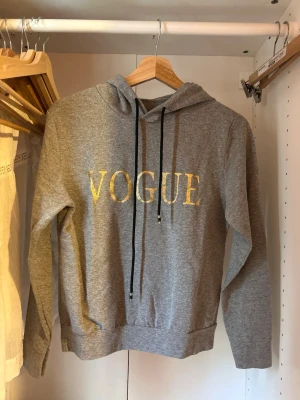 Grå hoodie med VOGUE-tryck - Snygg grå hoodie med guldigt VOGUE-tryck på bröstet och svarta dragsnören i huvan. Klassisk passform med långa ärmar och ribbade muddar. Perfekt för en avslappnad och trendig stil.