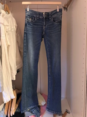 Blå bootcut jeans med dubbla knappar - Snygga blå bootcut jeans med dubbla knappar i midjan och klassiska fem fickor. Jeansen har en lätt tvättad look och kontrastsömmar. Perfekta för dig som gillar en retrostil med utsvängda ben. Materialet är troligtvis bomullsdenim.