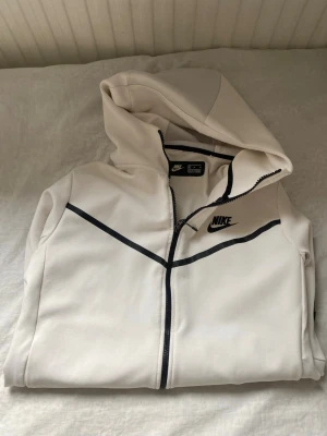 Vit Nike hoodie med dragkedja - Säljer en stilren vit hoodie från Nike med svart dragkedja och svarta detaljer. Hoodien har huva, två fickor framtill och Nike-logga på bröstet. Tillverkad i mjukt material som känns skönt mot huden. Perfekt för dig som gillar sportig och clean stil. Pris kqn diskuteras 