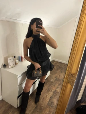 Svart omlottklänning med volang - Säljer en svart kort omlottklänning med volangkant och hög hals. Klänningen är ärmlös och har en flowy passform som ger en snygg siluett. Perfekt för dig som vill ha en trendig och chic look.