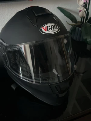 Svart Moped hjälm från Vcan - Säljer en svart integralhjälm från Vcan med klart visir. Hjälmen har ventilationsöppningar och mjuk vadderad insidq. Perfekt för nya moped användare och säker hjälm. Jag har själv inte haft moped så den har bara använts när jag åkt med mina kompisar. Aldrig tappad eller skadad! Storlek är YM 49-50cm (omkretsen av ditt huvud)