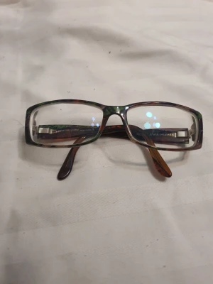 Glasögon med mönstrad båge - Säljer ett par rektangulära glasögon i brunt och grönt mönster. Bågarna har en stilren design och passar dig som vill ha en accessoar som sticker ut lite. Perfekt för att komplettera din look.Glasögon Johann von Goisern.