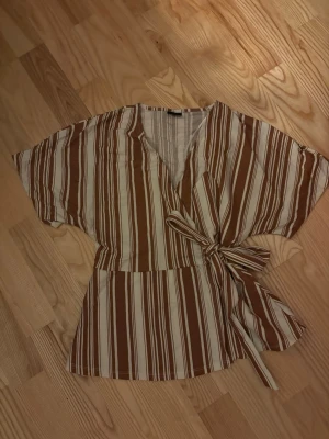 Randig, brun, knyttopp m. rosett - Säljer en brun och vit randig omlottopp med korta ärmar och knytband i midjan. Toppen har v-ringning och ett avslappnat fall, perfekt för en chill och trendig look. Materialet känns mjukt och lätt, troligen bomull eller viskos.