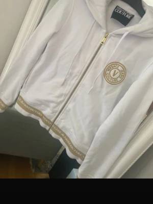 Vit hoodie från Versace Jeans Couture - Snygg vit hoodie från Versace Jeans Couture med guldfärgade detaljer och dragkedja framtill. Märkets logga syns tydligt på bröstet och längs nederkanten. Hoodien har huva med dragsko och är tillverkad i mjukt material för en bekväm känsla.