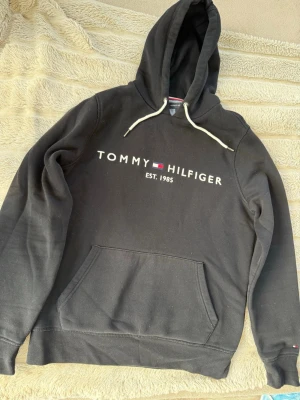 Svart hoodie från Tommy Hilfiger - Svart hoodie från Tommy Hilfiger med vit logga och text 'EST. 1985' på bröstet. Klassisk känguruficka framtill och vita dragsnören i huvan. Tillverkad i mjukt bomullsmaterial, perfekt för en avslappnad och stilren look.