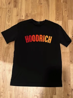 Svart HOODRICH t-shirt - Svart t-shirt från HOODRICH med broderad logga i rött, orange och gult framtill. Klassisk passform med rund hals och korta ärmar.