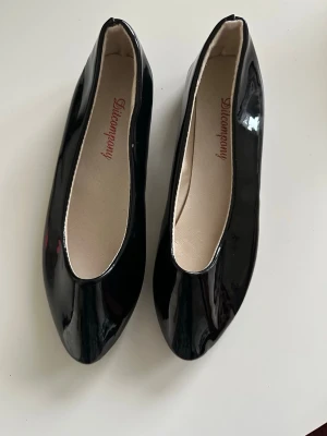 Svarta lackade ballerinaskor, storlek 38 - Säljer ett par stilrena svarta ballerinaskor från Discompany med blank lackfinish och rundad tå. Skorna har ljus innersula och klassisk form som passar till många olika outfits. Perfekt för dig som gillar enkel och elegant stil.