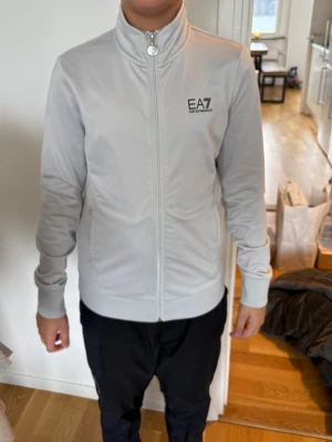 Vit ziptröja från EA7 Emporio Armani - Snygg vit ziptröja från EA7 Emporio Armani med hög krage och dragkedja hela vägen. Logga på bröstet och stort tryck på ryggen. Tillverkad i ett mjukt och stretchigt material som ger en sportig look. Perfekt till både jeans och träningsbyxor.