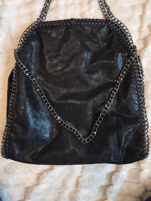 Svart axelväska med kedjedetaljer - Cool svart axelväska med glansig finish och kraftiga silverfärgade kedjor som axelrem och runt kanterna. Väskan har en mjuk, slouchy form och ger en edgy vibe till din outfit. Perfekt för dig som gillar statement-accessoarer.