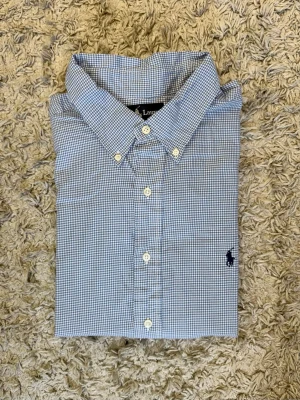 Blårutig skjorta från Ralph Lauren - Klassisk blårutig skjorta från Ralph Lauren med button-down krage och lång ärm. Skjortan har vita knappar framtill och den ikoniska broderade loggan på bröstet. Tillverkad i mjuk bomull som känns skön mot huden. Perfekt för en clean och stilren look.