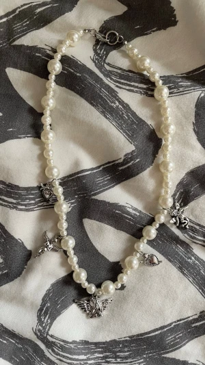 Pärlhalsband med silverberlocker - Snyggt halsband med vita pärlor och flera silverfärgade berlocker i form av ängel, hjärta och djur. Halsbandet har ett klassiskt pärlband med olika storlekar på pärlorna och ett metallspänne. Perfekt accessoar för att lyfta din outfit.