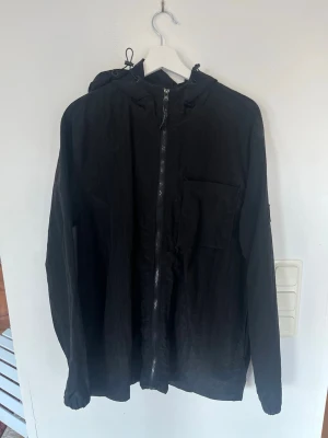 Lagency vindjacka  - Snygg svart Lagency vindjacka med dragkedja framtill och huva med dragsko. Jackan har en bröstficka och långa ärmar med resår vid ärmsluten. Perfekt för blåsiga dagar och enkel att matcha till olika outfits. Storleken är M men passar L bättre därav säljer jag den. Aldrig använd heller.