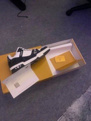 Louis Vuitton svartvita sneakers - Snygga svartvita Louis Vuitton sneakers med klassiska monogramdetaljer och vita stjärnor på sulan. Skorna har en låg silhuett, vita skosnören och är tillverkade i en mix av läder och textil. Perfekta för dig som vill sticka ut med exklusiv streetstyle.