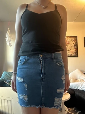 Blå jeanskjol med slitningar - Säljer en blå kort jeanskjol med slitna detaljer och rå kant nertill. Kjolen har klassisk femficksdesign, knapp och dragkedja framtill. Perfekt för dig som gillar en avslappnad och trendig stil.