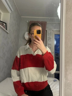 Randig stickad tröja med krage - Säljer en stickad tröja med breda röda och vita ränder. Tröjan har en klassisk krage och knäppning vid halsen. Långärmad och i mjukt material, perfekt för en avslappnad stil. Passar dig som gillar retrovibbar och sportig look.