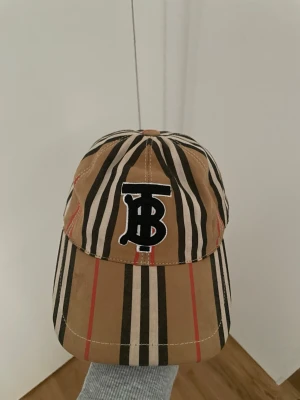 Randig keps från Burberry - Säljer en snygg keps från Burberry med klassiskt beige, svart, vitt och rött randigt mönster. Framtill sitter ett broderat svart och vitt TB-monogram. Kepsen har böjd skärm och justerbar rem bak. Perfekt för dig som gillar streetstyle och designeraccessoarer.