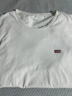 Vit t-shirt från Levi's med logga - Klassisk vit t-shirt från Levi's med rund halsringning och den ikoniska röda Levi's-loggan broderad på bröstet. Tillverkad i mjuk bomull för en skön känsla och enkel att matcha med allt. Perfekt basplagg för din garderob.