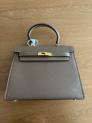 Begie handväska  - Väskan har en stilren, fyrkantig form och ett topphandtag. 