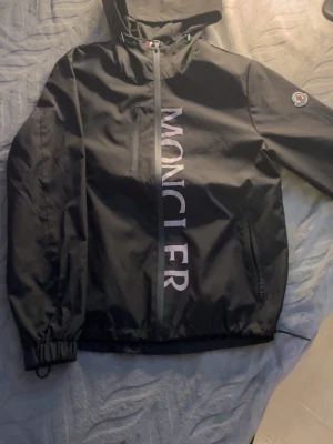 Svart vindjacka från Moncler - Snygg svart vindjacka från Moncler med stor vit logga längs dragkedjan framtill. Jackan har huva, två sidofickor med dragkedja och Moncler-märke på ärmen. Tillverkad i ett lätt och vattenavvisande material, perfekt för blåsiga dagar.