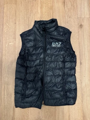Svart dunväst från EA7 Emporio Armani - Snygg svart dunväst från EA7 Emporio Armani med quiltad design och hög krage. Västen har dragkedja framtill och logga på bröstet. Perfekt att slänga över en hoodie eller tröja för extra värme och stil.