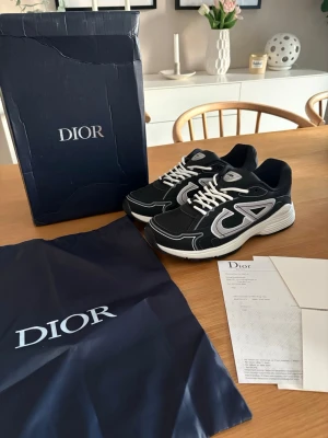 Svarta Dior sneakers med vita detaljer - Säljer ett par svarta Dior sneakers med vita och grå detaljer samt vita skosnören. Skorna har en chunky sula och tydlig Dior-logga på sidan och plösen. Materialet är en mix av mesh och läder för en sportig och exklusiv look. Kommer med originalkartong och dustbag.