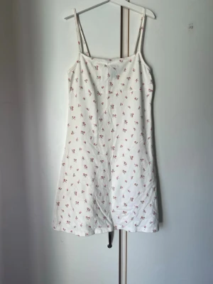 Vit blommig sommarklänning - Söt vit klänning med små rosa blommor och tunna axelband. Lätt och luftig modell som passar perfekt till varma dagar. Klänningen har en rak passform och är kort i längden. Perfekt för en avslappnad och trendig look.