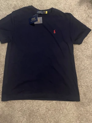 Marinblå t-shirt från Polo Ralph Lauren - STÅR L MEN PASSSR S.            Snygg marinblå t-shirt från Polo Ralph Lauren med klassisk röd logga broderad på bröstet. T-shirten har korta ärmar och är gjord i mjuk bomull för en skön känsla. Perfekt basplagg med stilren design och rund hals.