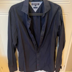 Mörkblå skjorta från Tommy Hilfiger - Snygg mörkblå skjorta från Tommy Hilfiger med klassisk krage och långa ärmar. Skjortan har knappar framtill och diskreta röda detaljer vid manschetten samt det lilla Hilfiger-flaggan broderad vid ärmslutet. Perfekt för en stilren look.