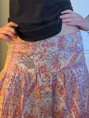 Färgglad långkjol  - Säljer en bohemisk långkjol med smock i midjan och färgstarkt paisleymönster i orange, rosa, lila och beige. Kjolen har en luftig och flowy passform, perfekt för varma dagar. Materialet känns lätt och mjukt mot huden.