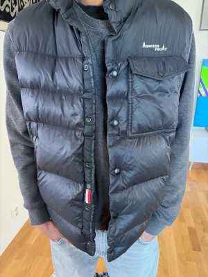 Svart dunväst från Moncler - Säljer nu min äkta Moncler × Fragment‑väst (verifierad via officiell QR‑kod på Moncler.com). Moncler Fragment väst för herr i svart färg. Yttermaterial i polyamid. Storlek 50, motsvarande Moncler storlek 3 enligt inneretikett.  Fint skick — jämn puff, ( men använd) Västen har quiltad, vadderad konstruktion med hög krage. Stängning med knappar framtill. Flera fickor, inklusive bröstficka med lock. Diskret Moncler-detalj framtill samt större Moncler Fragment-tryck på ryggen.  Prisförslag: 4500 kr 
