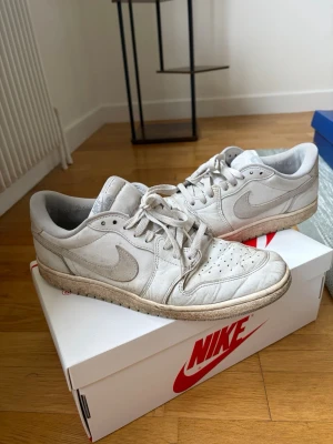 Nike Air Jordan 1 Low vita sneakers - Nike Air Jordan 1 Low sneakers i helvitt läder med klassisk swoosh på sidan och perforerad tåbox. Skorna har vita skosnören och en låg silhuett som ger en clean och stilren look. Perfekta för dig som gillar streetwear och tidlös design.