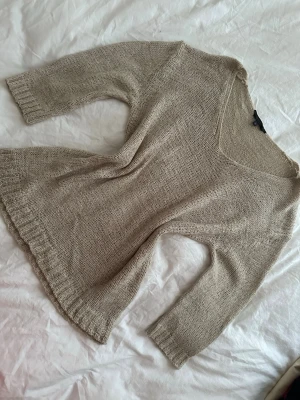 Beige stickad tröja med v-ringning - Beige stickad tröja med bred v-ringning och trekvartsärmar. Tröjan har en enkel, avslappnad passform och ribbstickade muddar vid ärmslut och nederkant. Perfekt för lager på lager och en stilren vardagslook.