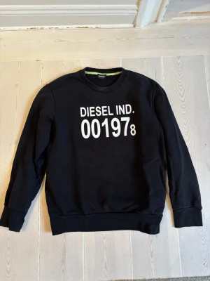 Blå sweatshirt från Diesel - Blå sweatshirt från Diesel med vit text 'DIESEL IND. 001978' på bröstet. Tröjan har rund hals, långa ärmar och ribbade muddar vid ärmslut och nederkant. Tillverkad i mjukt bomullsmaterial för en skön och avslappnad stil.