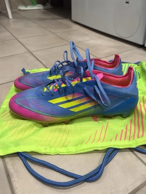 Adidas F50 - Säljer ett par Adidas F50 fotbollsskor i blått med neonrosa och gula detaljer. Skorna har snörning, låg profil och är tillverkade i syntetmaterial för bästa grepp på planen. Perfekta för dig som vill sticka ut på matchen! OBS Påse och box ingår👌