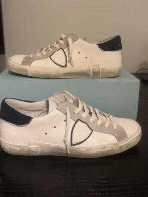 Vita Philippe Model sneakers med mockadetaljer - Snygga vita Philippe Model sneakers med grå mockadetaljer och mörkblå häl. Klassisk låg modell med vita snören och gummisula. Ikonisk sköldlogga på sidan och stilren design som passar till det mesta.