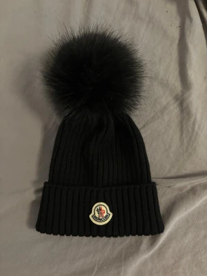 Svart Moncler mössa med pälsboll - Säljer en svart ribbstickad Moncler mössa med stor fluffig pälsboll på toppen och klassisk Moncler-logga framtill. Mössan är tillverkad i Italien och har en uppvikt kant för extra stilpoäng. Perfekt accessoar för kalla dagar.