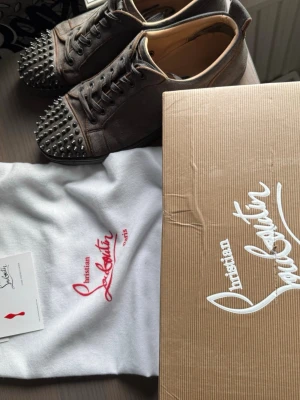 Christian Louboutin skor - Unika grå sneakers från Christian Louboutin i mocka med ikoniska röda sulor och coola svarta nitar på tån. Skorna har snörning och rund tå, samt exklusiv känsla med lyxiga material och detaljer. Perfekta för dig som vill sticka ut med din stil. Skriv vid minsta fundering! Storleken passar 44-46