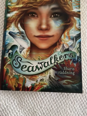 Seawalkers: Sharis räddning - En färgstark bok med fantasifullt omslag som visar en ung person, delfin och undervattensmiljö. 'Seawalkers: Sharis räddning' är andra delen i serien och handlar om äventyr på Blue Reef High, vänskap och mysterier i havet. Perfekt för dig som gillar spännande ungdomsböcker.