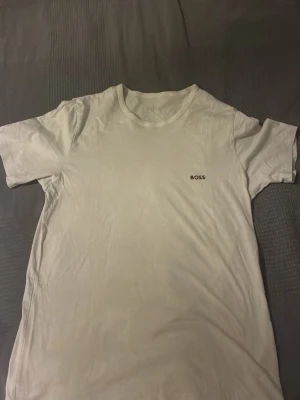 Vit t-shirt från BOSS - Säljer en stilren vit t-shirt från BOSS med diskret logga broderad på bröstet. Klassisk rund halsringning och korta ärmar. Perfekt basplagg i mjuk bomull som funkar till allt. Enkel och clean design för en modern look.