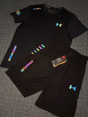 Svart träningsset från Under Armour - Säljer ett svart träningsset från Under Armour med t-shirt och shorts. Setet har färgglada loggor och textdetaljer i regnbågsfärger. Materialet är snabbtorkande och stretchigt, perfekt för träning eller gym. Snygg och sportig look med elastisk midja på shortsen.
