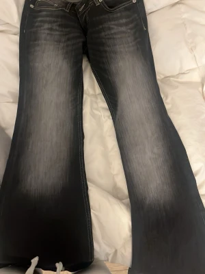 Lågmidjade svarta bootcut jeans med slitningar - Säljer ett par svarta bootcut jeans med snygga slitningar och ljusare partier framtill och baktill. Jeansen har klassisk femficksmodell, kontrastsömmar och är tillverkade i denim. Perfekta för dig som gillar en retro och avslappnad stil.