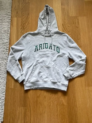 Grå Axel Arigato hoodie - Snygg grå hoodie från Arigato med grön text på bröstet. Klassisk modell med stor huva, dragsko och känguruficka framtill. Mjuk bomullsblandning och ribbade muddar vid ärmslut och midja. Perfekt för en avslappnad streetstyle-look. 